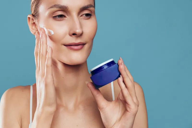 A woman with clear skin applies moisturizing cream to her face holding a blue jar against a plain blue background. Concept for skincare routine and beauty care.