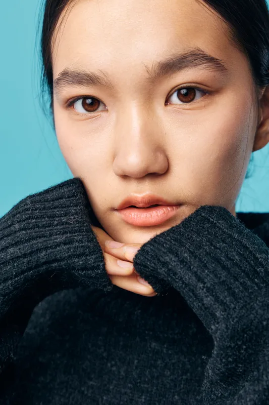 Woman with natural skin and dark hair wearing black sweater resting chin on hands with blue background. Closeup portrait showing calm expression and smooth complexion indoors.