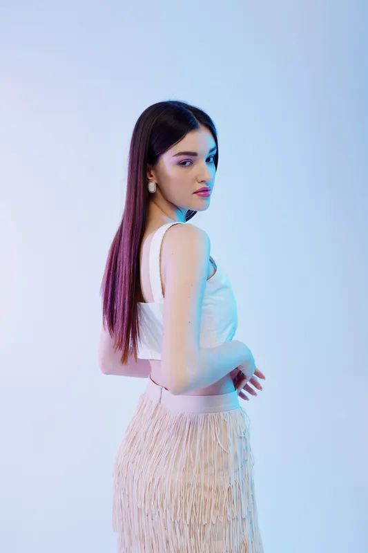 Young woman with long dark hair poses in a white top and fringed skirt. Studio shot with soft lighting on a light background. Concept of beauty fashion and style.