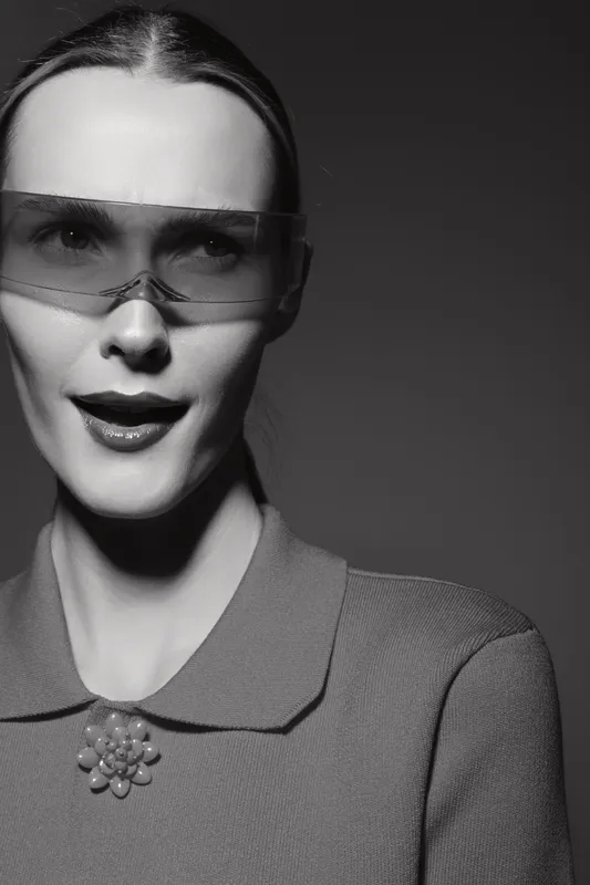 White woman with pale skin wearing futuristic sunglasses and vintage outfit poses in black and white studio portrait with moody lighting and confident expression.