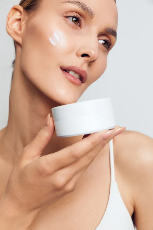 Young woman with clear skin holding white cream jar applying face moisturizer on cheek. Close-up portrait with soft lighting on light background. Concept of skincare and beauty care.