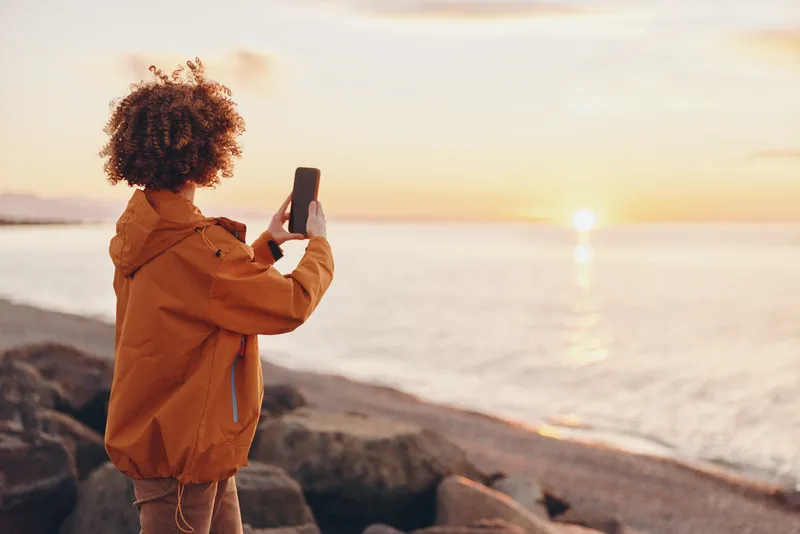 Woman with curly hair in a brown jacket holds a smartphone by the seaside at sunset, calm travel lifestyle, outdoor evening light, looking at screen, peaceful mood, during golden hour