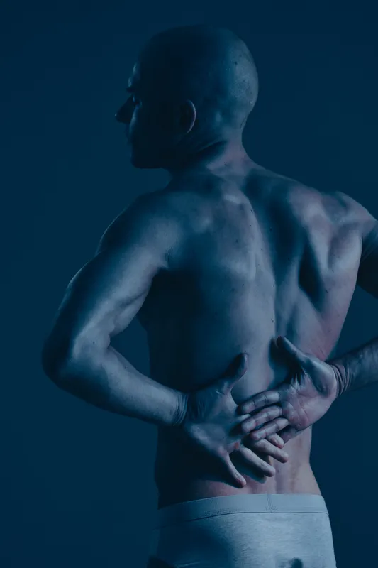A man with a muscular build experiencing back pain, portrayed in a dramatic blue light. The image captures the intensity of discomfort in a compelling manner.