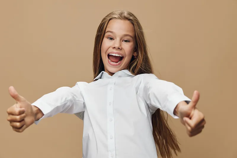 Happy girl with long hair smiling and showing thumbs up gesture wearing white shirt isolated on beige background for positive expression and approval.