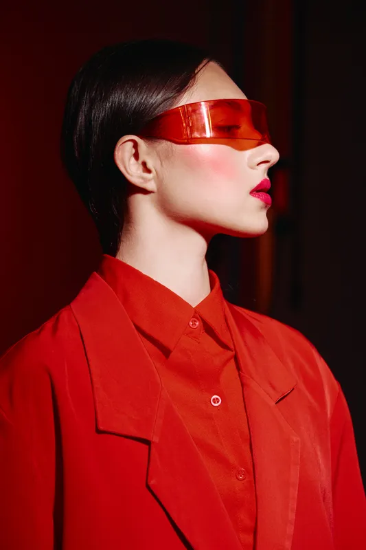 Profile portrait of a fashion model with vibrant red lipstick wearing futuristic orange visor and matching red stylish blazer and shirt in a dramatic lighting setup.