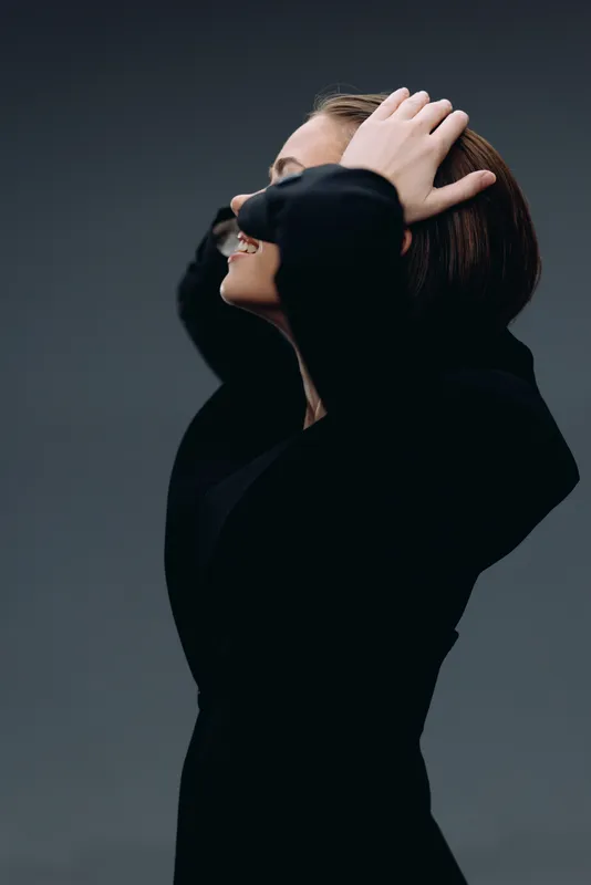 Profile of smiling woman in black dress with hands in hair on dark background