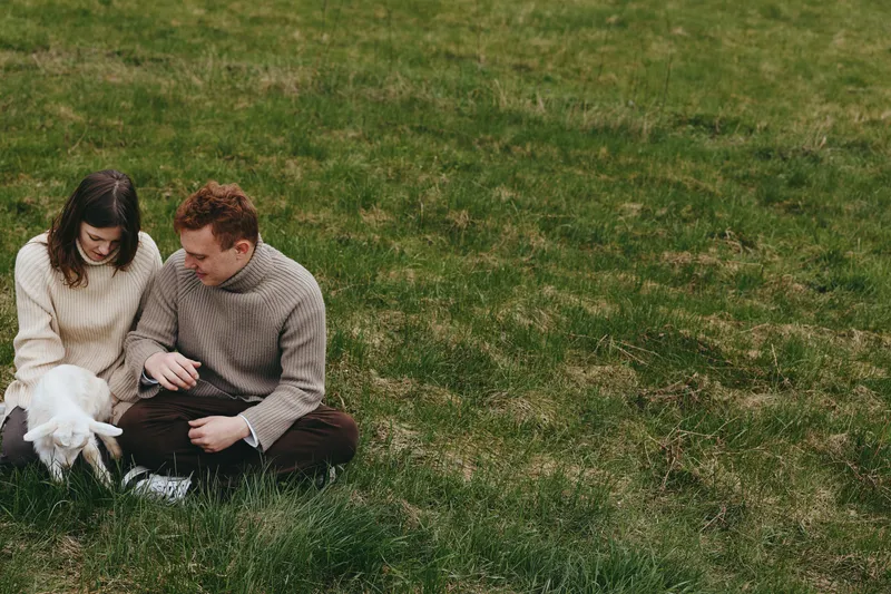 Cozy couple outdoors, relaxed moment with a playful goat kid beside them, soft sweaters, sunlit grass, gentle breeze, natural colors, casual companionship in a peaceful field