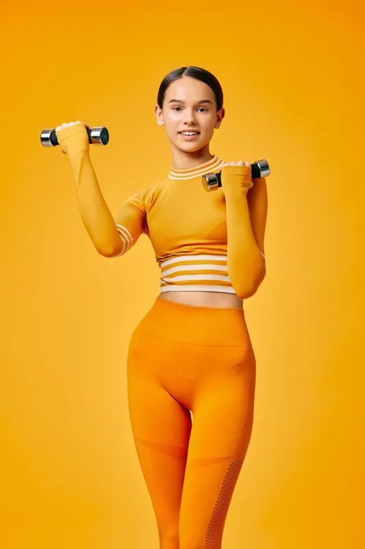 Young athletic woman in matching orange fitness set lifting small dumbbells in a bright studio, confident posture, toned silhouette, modern activewear, energetic mood.