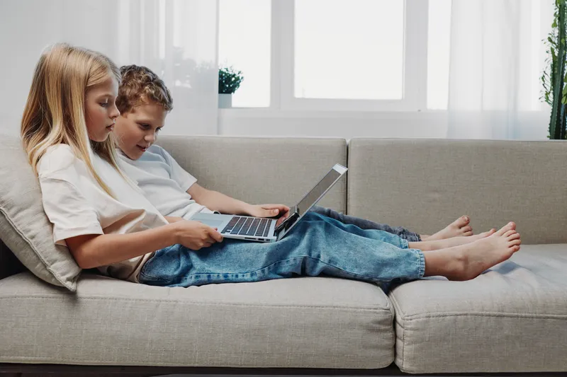 Two children sitting on a sofa using a laptop together in a bright modern living room, focused and engaged in digital learning or entertainment.