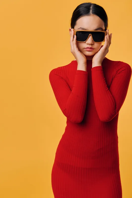 Woman in red dress posing with sunglasses on yellow background. Fashion, style and elegance with confident look and modern clothing in studio portrait photo.