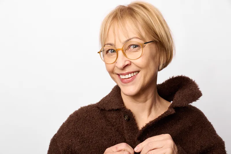 Smiling senior woman portrait with glasses and blond hair, wearing a dark fur collar coat on a white studio background, expressing smile, fashion, and confidence, relaxed pose.