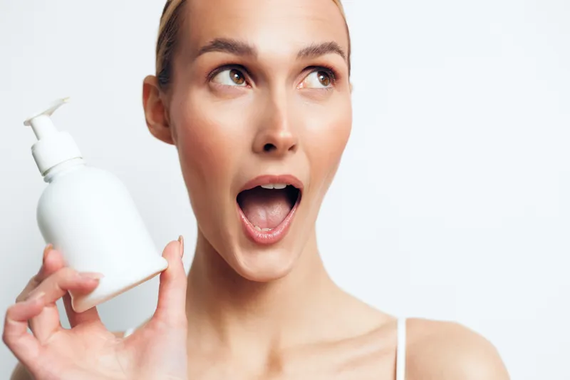 A woman with clear skin holds a pump bottle, eyes wide, mouth open in astonishment. Bright studio lighting highlights skincare routine and the moment of discovery.