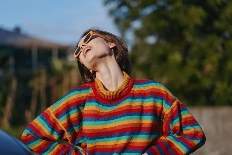 young woman in her 20s wearing a striped sweater and yellow sunglasses, head tilt and smiling outdoor in a sunny urban park, casual outfit and carefree lifestyle portrait