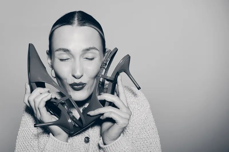 Young caucasian woman with light skin holds two high heel shoes near her face, eyes closed with joyful expression, studio shot.