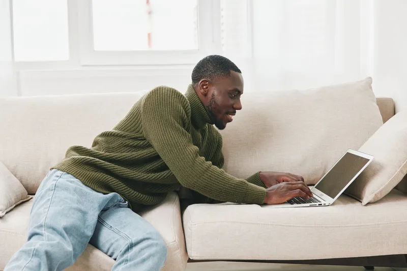 Relaxed young man working on laptop in cozy living room, wearing a green sweater and jeans, smiling, home office concept, modern interior style