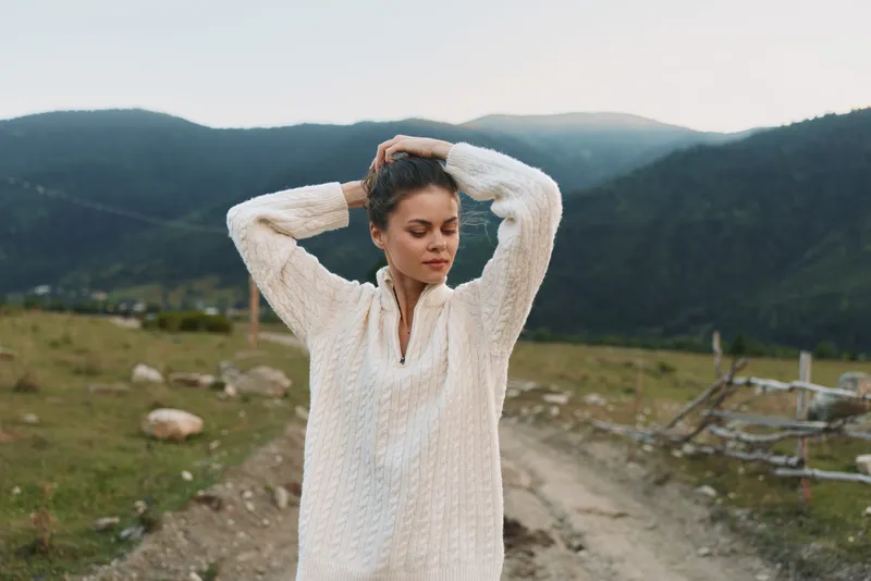 Smiling woman wearing a soft cream sweater stands along a dirt track surrounded by distant mountains, conveying a relaxed outdoor lifestyle and tranquil nature and warmth.