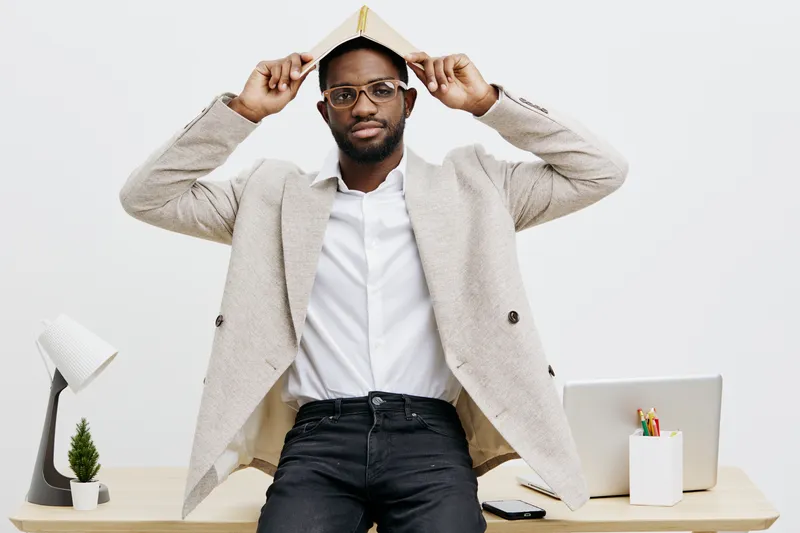 Professional portrait of a stylish African American man wearing glasses, beige blazer and white shirt, holding a hat on his head, seated at a modern white desk with a computer and stationery, neutral 