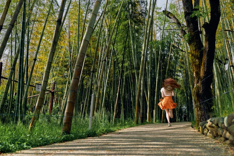 Girl running in a bamboo forest on a shaded path, orange dress flowing in motion through nature. Back view captures carefree movement, sunlight and tall stalks along a tranquil trail.