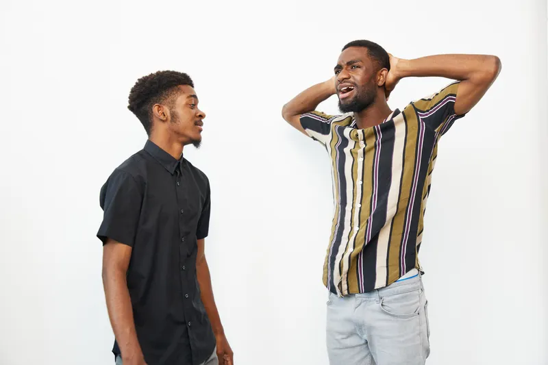 Two black men expressing a range of emotions, one laughing joyfully while the other listens in a vibrant, modern setting. Perfect for depicting friendship and joy.