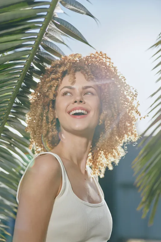 smiling woman with curly hair enjoying sunny tropical outdoors wearing white tank top surrounded by green palm leaves expressing happiness and warmth