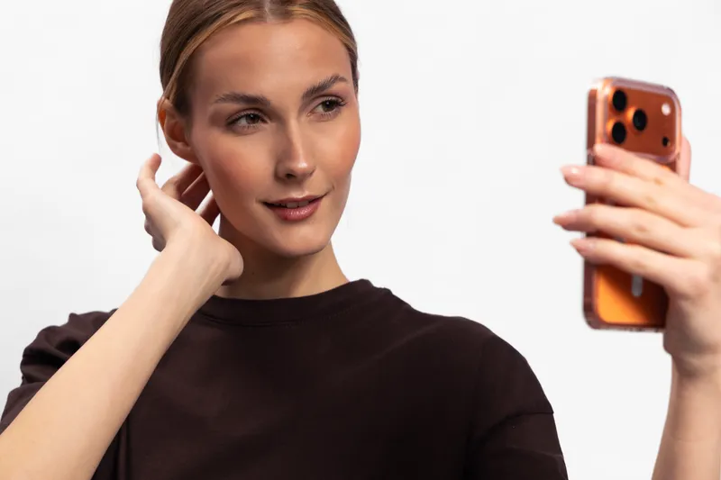 Young woman with light skin and tied hair takes selfie with smartphone, wearing dark shirt. Indoor studio photo with soft lighting, concept of communication, technology, and social media.