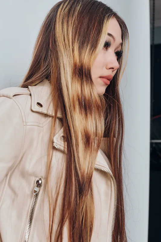 Portrait of a young woman with long straight hair wearing a beige leather jacket looking aside against a neutral background in casual style.