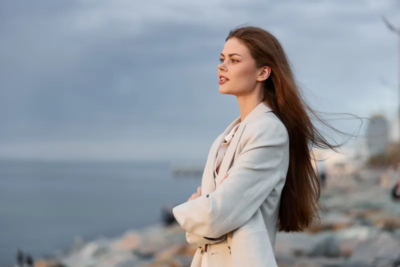 Woman in beige coat standing thoughtfully by rocky seaside with windy long hair and cloudy sky background, contemplative mood, autumn style fashion