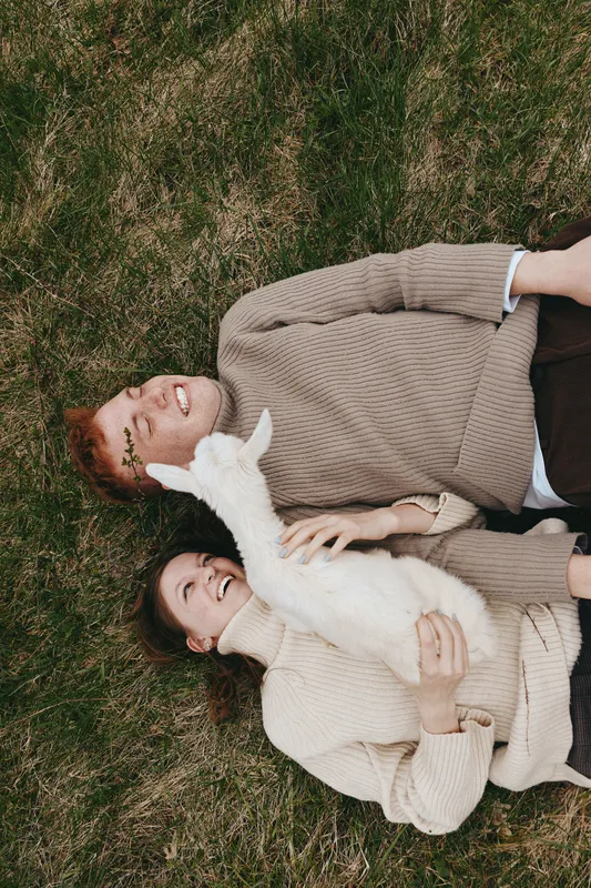 Cheerful couple lying on grass outdoors, smiling as a goat kid nuzzles them, cozy sweaters, sunlit breeze, relaxed lifestyle moment, natural colors, countryside vibes, candid connection