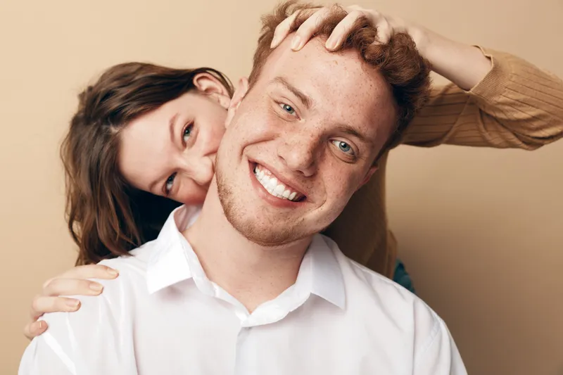 Happy young couple with smiling man in white shirt and woman playfully hugging him from behind on beige background. Concept of love, friendship, joy, and positive emotions.