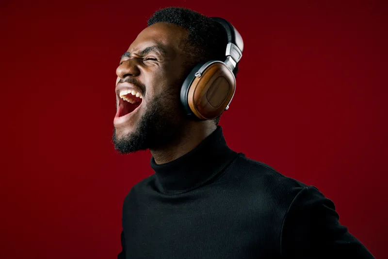 Enthusiastic Black man enjoying music with headphones on a vibrant red background, expressing joy and excitement through music, stylish attire