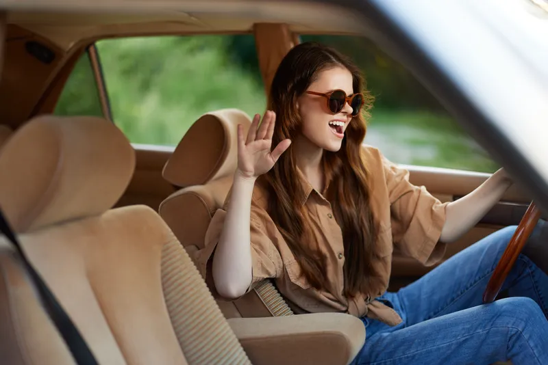 Young woman enjoying a road trip in a stylish car, waving and singing joyfully, dressed casually in a beige shirt and jeans, capturing a carefree summer vibe.