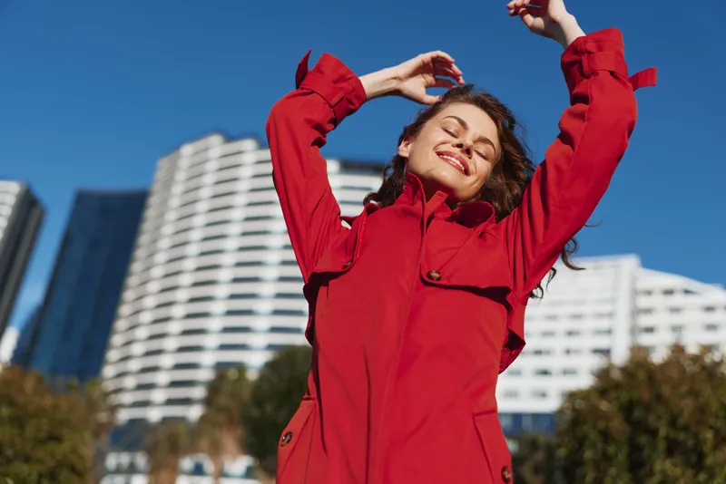 Young woman in a red coat stretches her arms upward with closed eyes and a joyful smile against a blurred urban background on a sunny day.