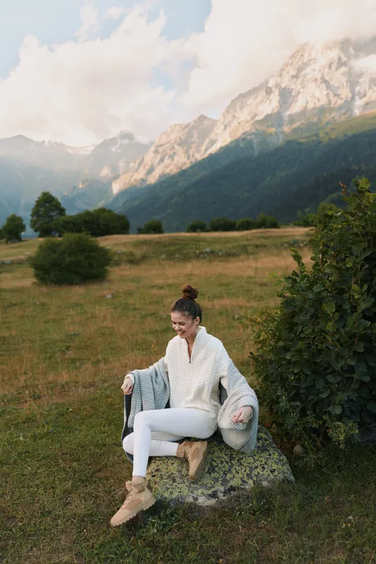 Woman outdoors in a mountain meadow, wearing a cozy sweater, sitting on a rock, relaxed pose, soft natural light, enjoying the fresh breeze, lifestyle moment