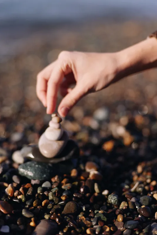Hand stacks stones and pebbles on a beach, creating balance and calm in nature. A mindful zen moment of outdoor sculpture and simple shore art amid textured gravel and water