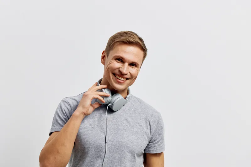 Smiling young man wearing casual gray t-shirt with headphones around neck, holding one hand near ear, looking at camera, cheerful expression, modern studio background, lifestyle concept, positive mood