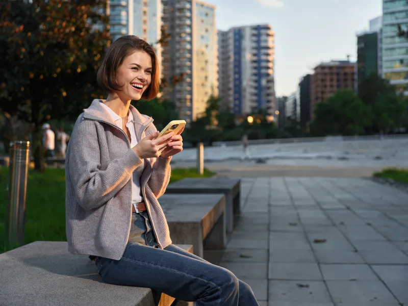 A cheerful young woman sits on a concrete bench in a modern city park, texting on a smartphone with a bright smile, exuding casual confidence and connectivity.