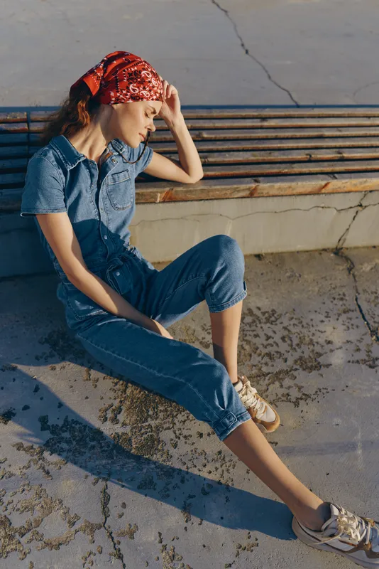 Lifestyle woman in modern boho-western denim street style wearing bandana sitting thoughtfully on urban bench in warm natural light with film color effects.