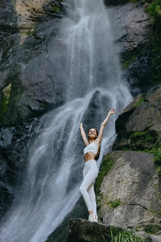Smiling woman enjoying waterfall nature, wearing white sport outfit, standing on rock with arms raised in joyful pose near flowing water and lush greenery.
