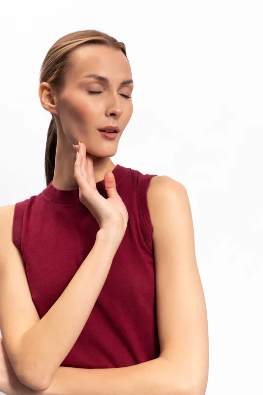 Young woman in sleeveless red top with closed eyes and hand near face poses on white background. Concept of beauty, fashion, elegance, and natural expression in studio lighting.