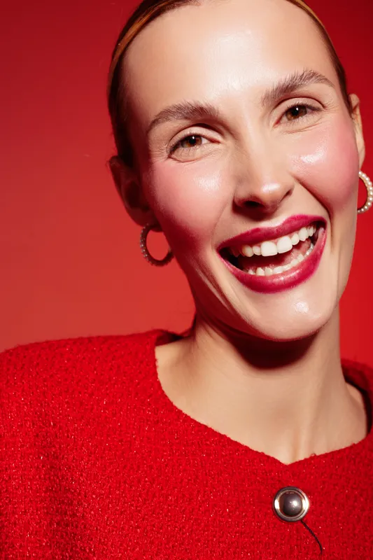 Smiling woman with red lipstick and hoop earrings wears red sweater against red background. Close-up portrait with natural skin glow and joyful expression. Concept of beauty and happiness.