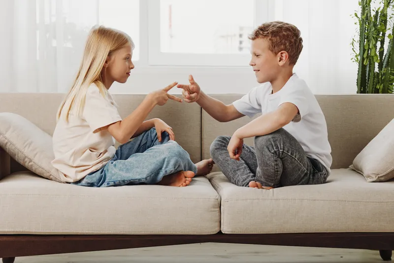 Two children playing rock paper scissors on a beige couch in a bright living room, enjoying a relaxed moment together. Concept of childhood, friendship, and indoor play.