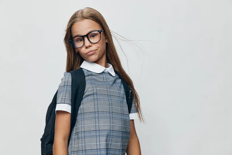 Girl child with long hair and glasses wearing backpack and plaid dress on white background for school, education and childhood concept with casual style and serious expression