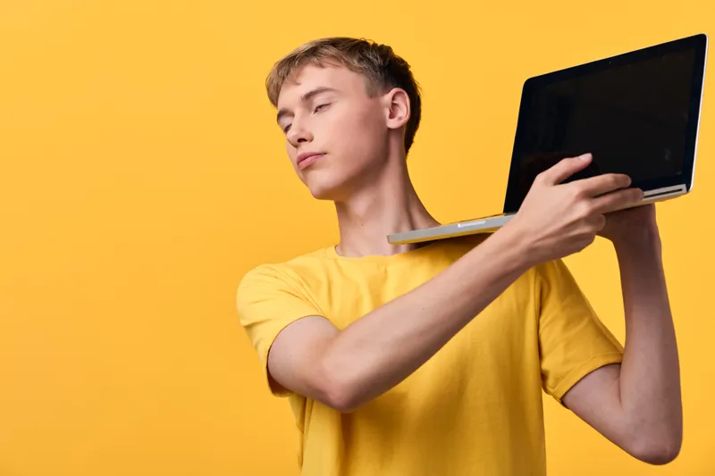 Man in a yellow t shirt balances a laptop on his shoulder against a bright yellow backdrop, showcasing portable tech, mobility and modern work style