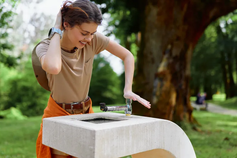 Woman drinking water from a concrete fountain in a park, outdoor candid portrait with casual clothes and smile, young adult staying hydrated during a sunny nature walk.