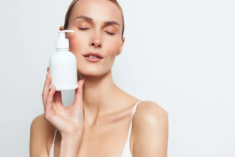 Young woman with clear skin holds white bottle with pump near face, eyes closed, expressing freshness and skincare routine on light background, minimalistic beauty concept.