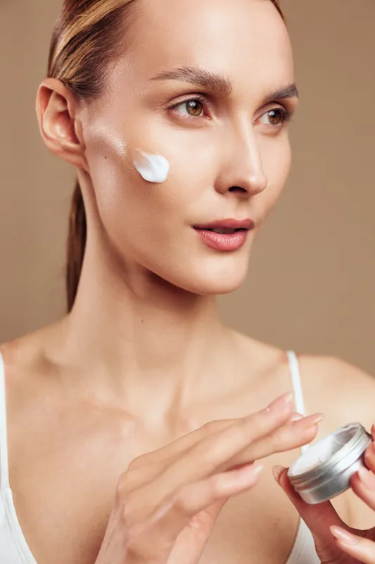 Woman skincare closeup, applying cream under eye, holding jar with applicator swab, calm expression, soft daylight, smooth face skin, beauty treatment
