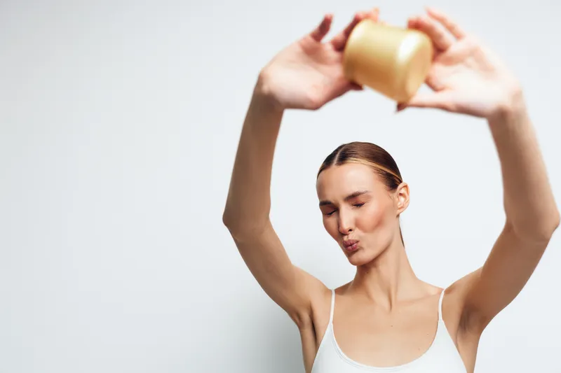 Young woman with closed eyes holds gold party cup above head celebrating with joyful expression isolated on white background in casual white tank top.