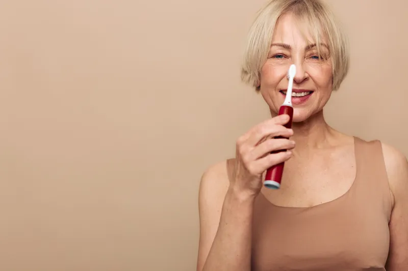 Smiling mature woman with short blonde hair holding electric toothbrush near face on beige background. Concept of oral hygiene, dental care, and daily routine for healthy teeth.