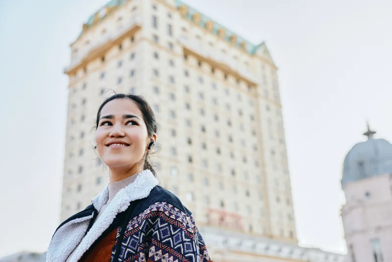 Urban woman in a stylish jacket smiles near a tall building, outdoor cityscape with architecture, showcasing fashion, confidence and travel vibe in a modern urban setting