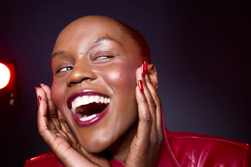 Close-up of joyful dark skinned African woman with shiny skin and red nails smiling widely against dark background. Concept of happiness, beauty, and self-confidence in studio lighting.